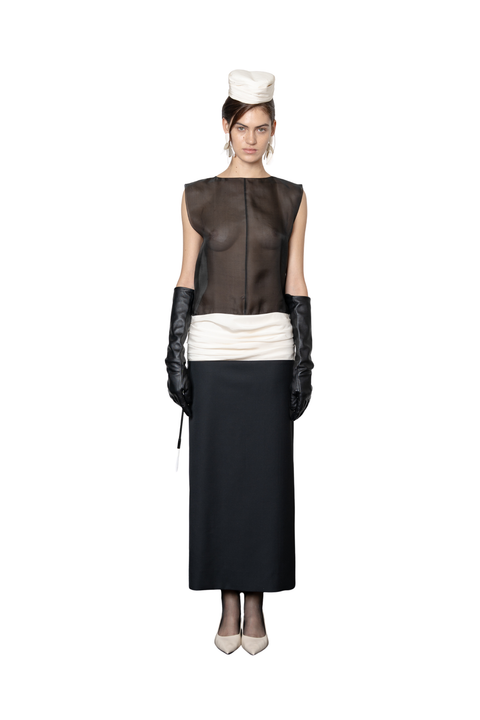A model stands against a white background wearing a black and white long dress consisting of a sheer black boat neck sleeveless top, a white gathered drop waistband, a long black skirt, black gloves, and a white hat.