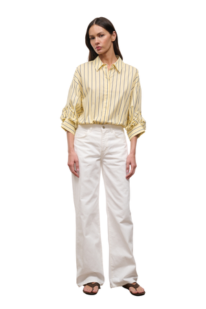 A woman modeling a pale yellow and white vertically striped button-down shirt with rolled sleeves, paired with white wide-leg trousers and brown sandals, against a plain white background.