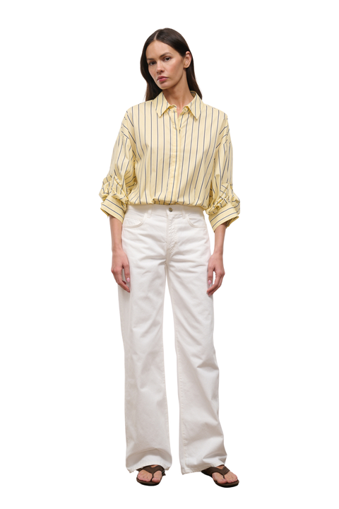 A woman modeling a pale yellow and white vertically striped button-down shirt with rolled sleeves, paired with white wide-leg trousers and brown sandals, against a plain white background.