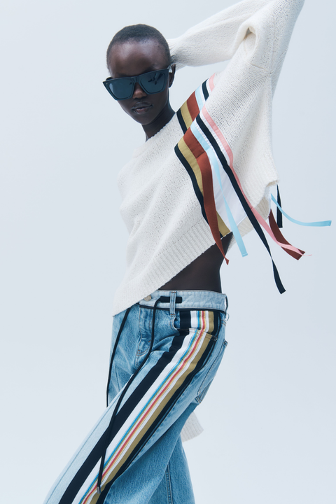Person wearing a white sweater with colorful ribbon stripes with fringe and blue jeans on a light gray background