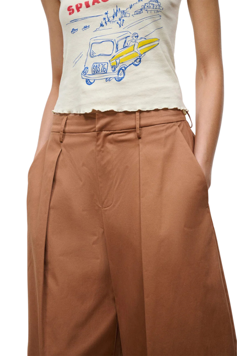 A close-up of a model wearing high-waisted, wide-leg brown trousers with pleats and a cream-colored graphic tank top.