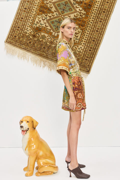 Side-facing woman wearing a multicolor, printed, mini shirtdress that features a point collar and self-tie belt, paired with brown mules. 