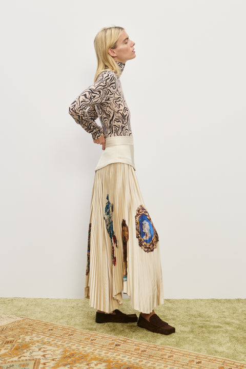 Side-facing woman wearing a cream midi skirt with an asymmetrical corseted waist and a pleated skirt with an asymmetrical hem line and colorful icons printed on the skirt, paired with a printed long sleeve top and brown loafers. 