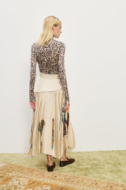 Back-facing woman wearing a cream midi skirt with an asymmetrical corseted waist and a pleated skirt with an asymmetrical hem line and colorful icons printed on the skirt, paired with a printed long sleeve top and brown loafers. 