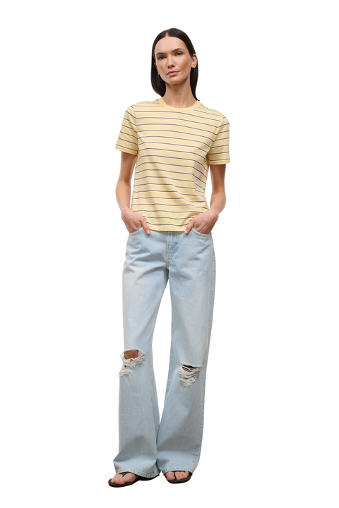 A woman modeling a short-sleeve crewneck t-shirt with thin yellow horizontal stripes on a light beige background, paired with light-wash distressed jeans and dark sandals, against a plain white studio background.