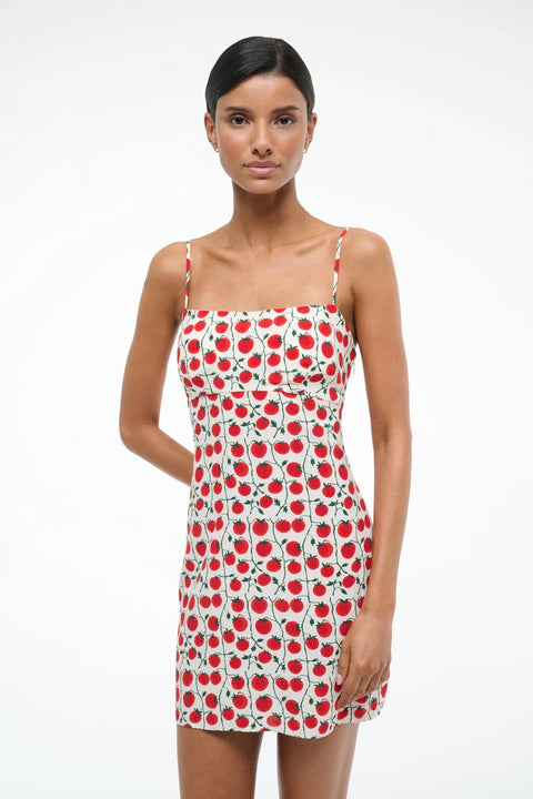 Close up of a woman wearing a white mini dress that features spaghetti straps, and square neck, and a red and green tomato pattern. 