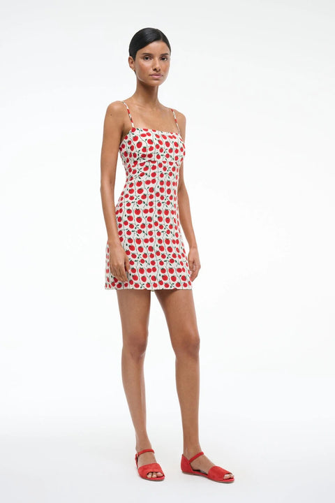Side view of a woman wearing a white mini dress that features spaghetti straps, and square neck, and a red and green tomato pattern, paired with red sandals. 