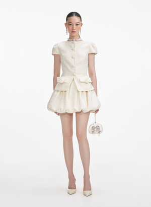 Woman wears cream boucle mini dress with bubble skirt hem and bow details.