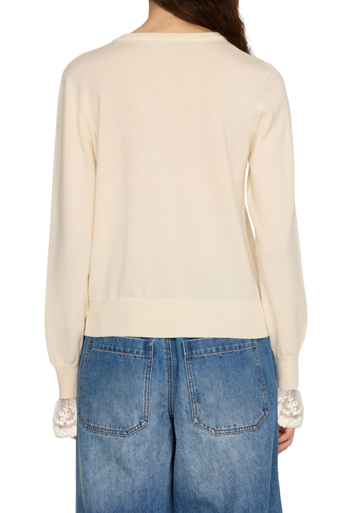 Back view of a woman wearing a cream-colored long-sleeve knit sweater with lace cuff details and blue jeans, against a plain white background.