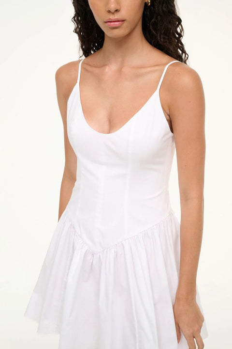 Close up of a woman wearing a white mini dress that features spaghetti straps and a scoop neck, a fitted bodice, and an A-line skirt.
