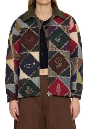 A model wearing a vintage-style patchwork quilted jacket with embroidered diamond motifs, styled over a dark top and brown trousers.