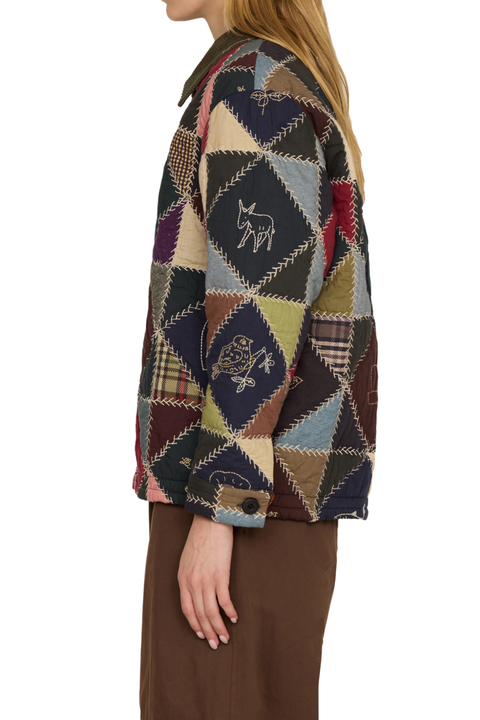A woman shown from the side wearing a patchwork quilted jacket with embroidered cow motifs and brown pants, against a plain white background.