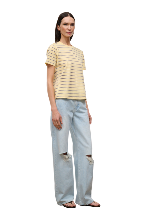 A woman modeling a short-sleeve crewneck t-shirt with thin yellow horizontal stripes on a cream base, paired with light-wash distressed jeans and brown sandals, set against a plain white background.