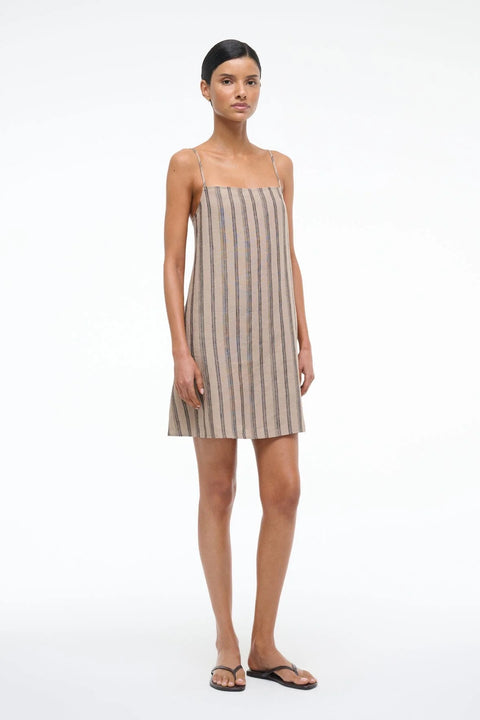 Side view of a woman wearing a spaghetti strap, shift mini dress crafted from a brown striped linen, paired wit brown sandals. 
