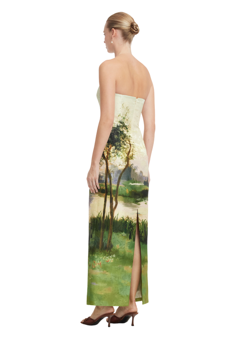 A strapless floor-length gown shown from the back on a model, featuring an artistic landscape print with trees, grass, and water in soft green, beige, and blue tones.