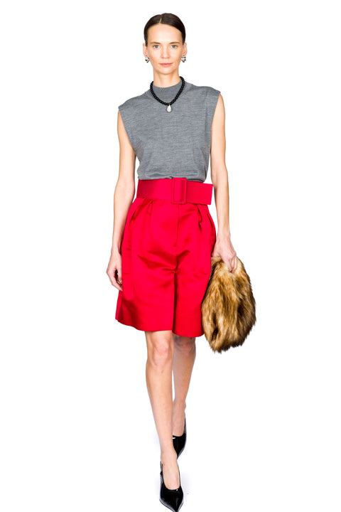 Woman walking forward wearing a gray sleeveless top and red shorts, holding a fur bag on a white background