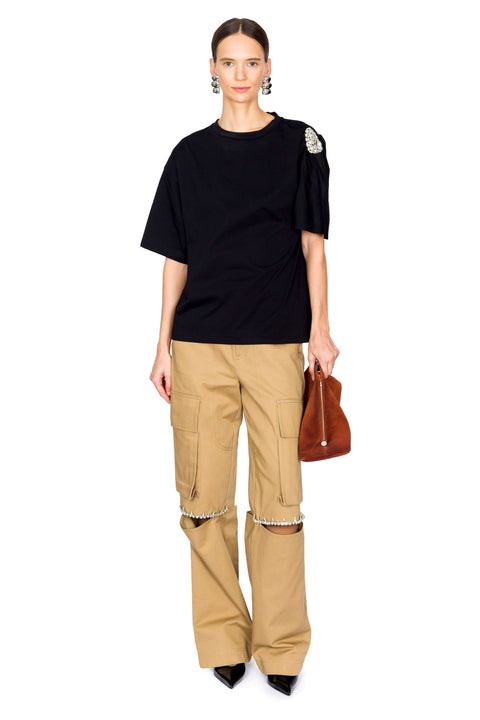Woman wearing a t-shirt with crystal embellishment on one shoulder and beige cargo pants holding a brown bag on a white background
