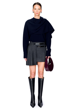 Woman wearing a navy blue sweater, gray pleated skirt, and black knee-high boots on a white background