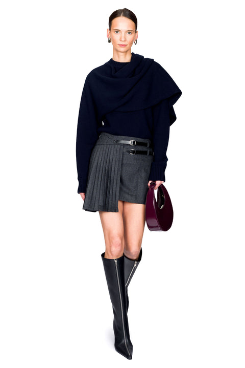 Woman wearing a dark blue sweater, gray pleated skirt, red handbag, and black knee-high boots on a white background