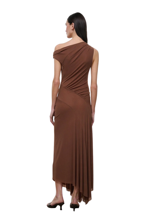 A woman modeling a brown one-shoulder draped maxi dress from the back, paired with black high heels, against a plain white background.
