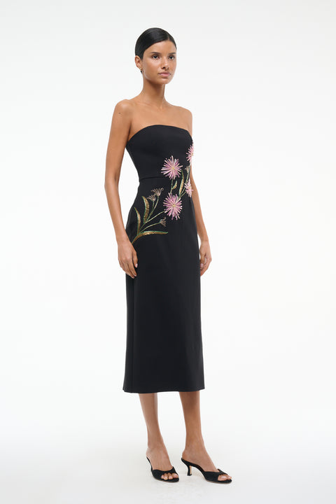 Side view of a woman wearing black, strapless midi dress that features pink dahlia embellishments across the bodice, paired with black heels. 
