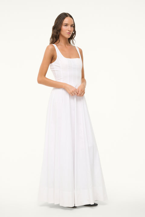 Side view of a woman wearing a sleeveless, white maxi dress that features a square neck, corset style paneling, and A-line skirt. 