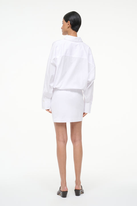 Back-facing woman wearing a long sleeve, white, mini shirt dress, paired with brown mules. 