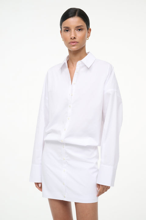 Close up of a woman wearing a long sleeve, white, mini shirt dress, paired with brown mules. 
