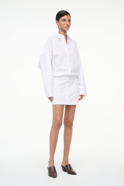 Side-facing woman wearing a long sleeve, white, mini shirt dress, paired with brown mules. 
