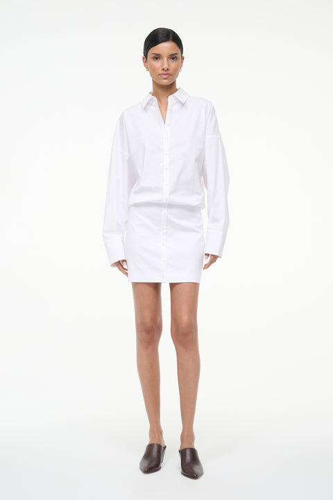 Woman wearing a long sleeve, white, mini shirt dress, paired with brown mules. 