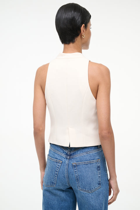 Back-facing woman wearing a cream button up vest with blue jeans. 