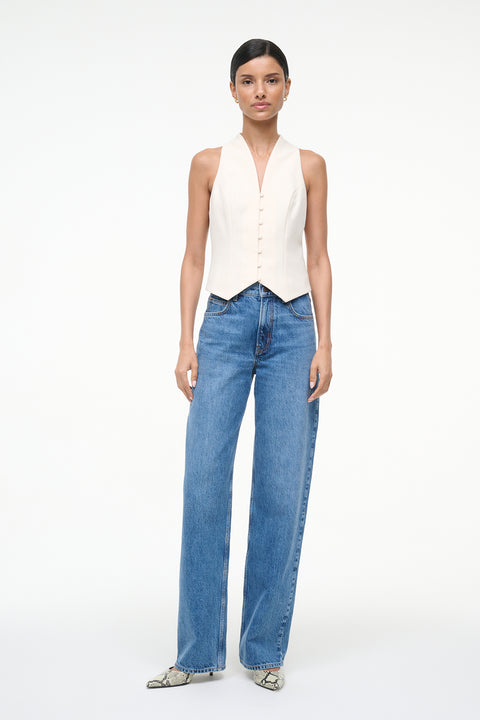 Woman wearing a cream button down vest that features a V neckline, paired with blue jeans. 