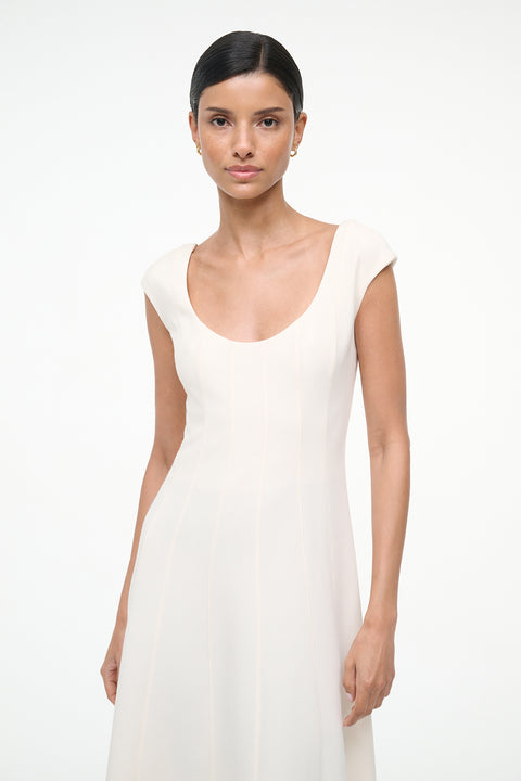Close up of a woman wearing a cream, A-line midi dress that features a scoop neck, cap sleeves, and princess seams at the waist. 