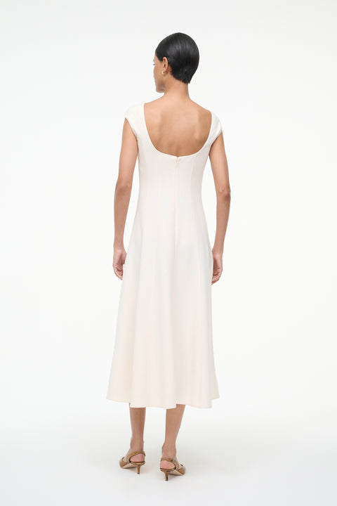 Back-facing woman wearing a cream, A-line midi dress that features a scoop back, cap sleeves, and princess seams at the waist, paired with kitten heels. 