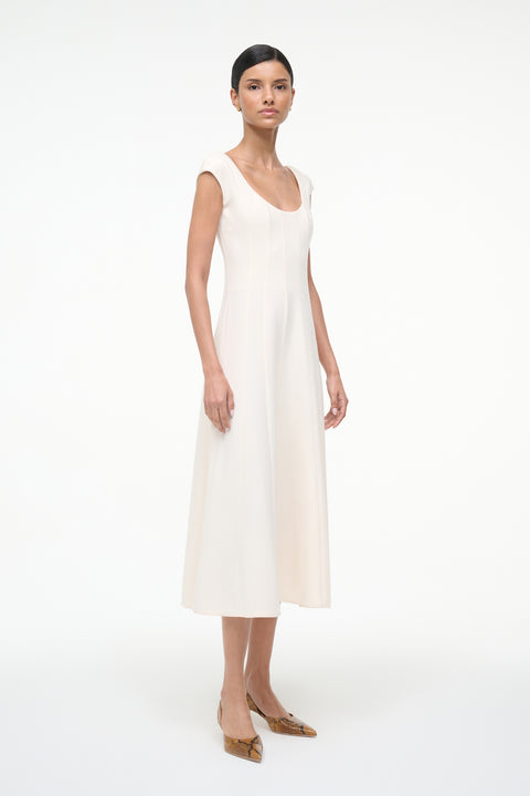 Side-facing woman wearing a cream, A-line midi dress that features a scoop neck, cap sleeves, and princess seams at the waist, paired with kitten heels. 