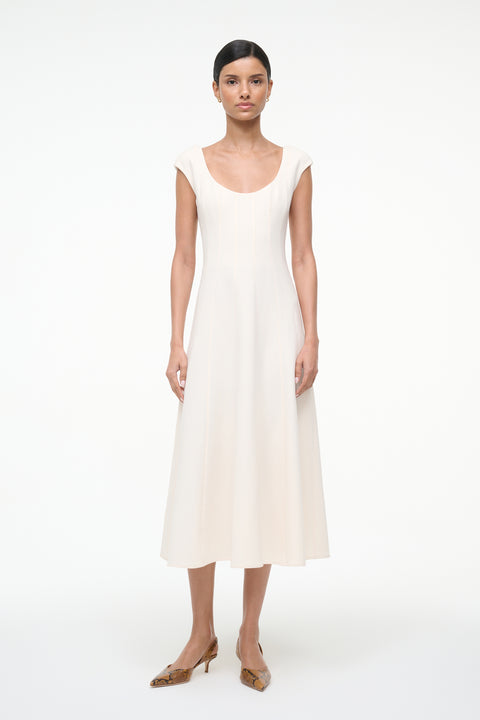 Woman wearing a cream, A-line midi dress that features a scoop neck, cap sleeves, and princess seams at the waist, paired with kitten heels. 