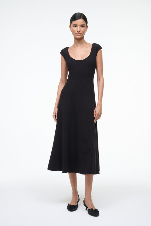Woman wearing a black, A-line, midi dress that features a scoop neck, princess seams at the waist, and cap sleeves, paired with black kitten heels. 