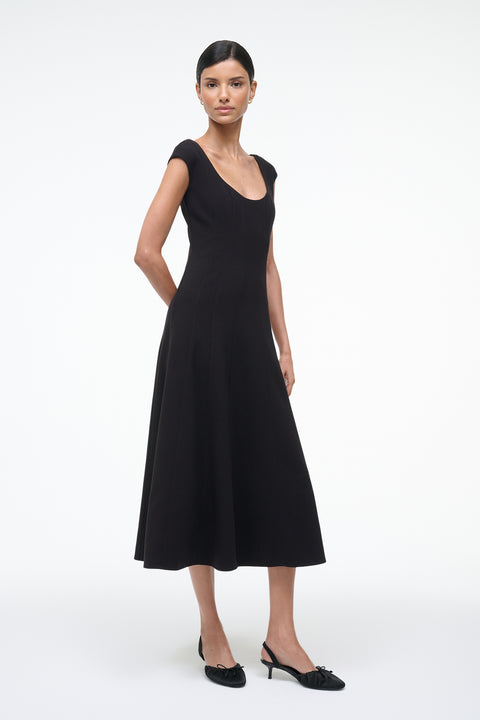 Side-facing woman wearing a black, A-line, midi dress that features a scoop neck, princess seams at the waist, and cap sleeves, paired with black kitten heels. 