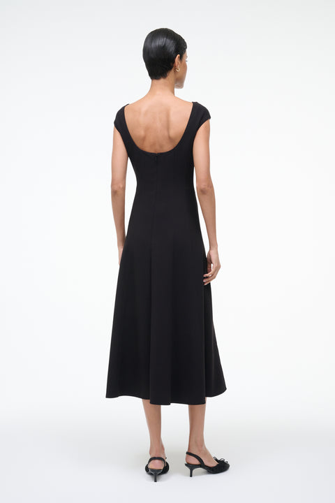 Back-facing woman wearing a black, A-line, midi dress that features a scoop back, princess seams at the waist, and cap sleeves, paired with black kitten heels. 