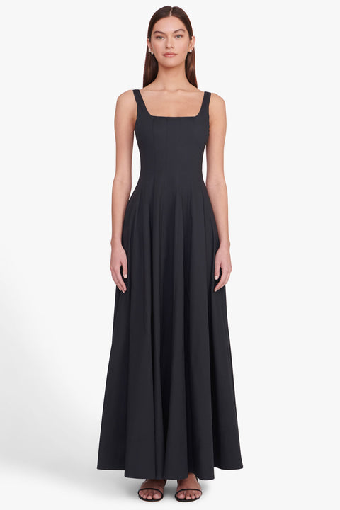 Woman wearing a sleeveless, black maxi dress that features a square neck, corset style paneling, and A-line skirt, paired with black sandals.