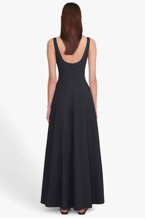 Back view of a woman wearing a sleeveless, black maxi dress that features a scoop back, corset style bodice, and A-line skirt, paired with black sandals. 
