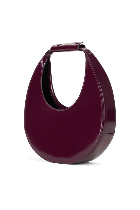 Side angle of a deep maroon, patent leather top handle bag inspired by the shape of a crescent moon. 