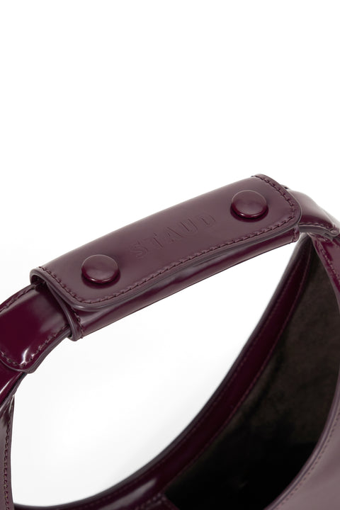 A deep maroon, patent leather top handle bag inspired by the shape of a crescent moon with a snap leather handle.