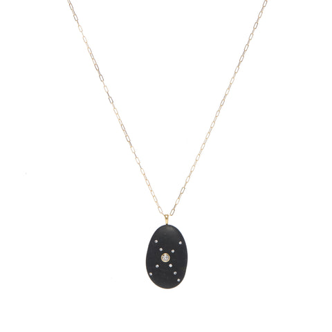 A black oval-shaped stone pendant that features a round diamond in the center of a crisscross pattern of smaller round diamonds, suspended on a gold paperclip chain. 