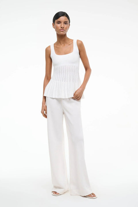 Woman wearing a white knit tank top that features a square neck and sheer, pleated bodice, paired with white pants and sandals. 