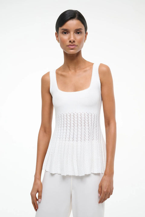 Close up of a woman wearing a white knit tank top that features a square neck and sheer, pleated bodice, paired with white pants. 