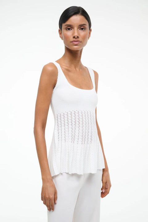 Side view of a woman wearing a white knit tank top with a square neck and sheer, pleated bodice, paired with white pants. 