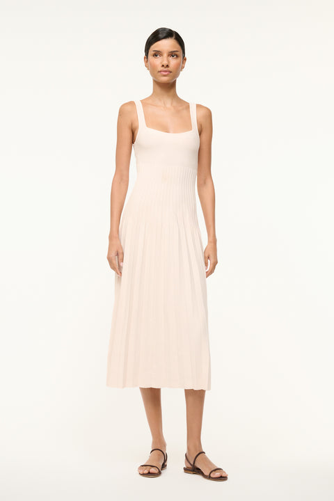 Woman wearing a cream knit midi dress that features a ribbed bodice and pleated skirt, paired with brown sandals. 