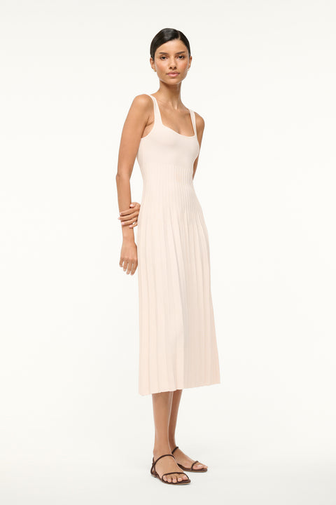 Side view of a woman wearing a cream knit midi dress that features a ribbed bodice and pleated skirt, paired with brown sandals. 