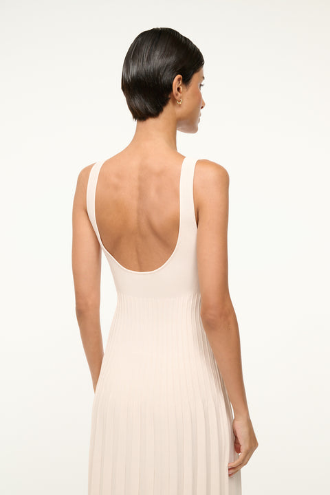 Close up back view of a woman wearing a cream knit midi dress that features a low scoop back, a ribbed bodice, and a pleated skirt. 